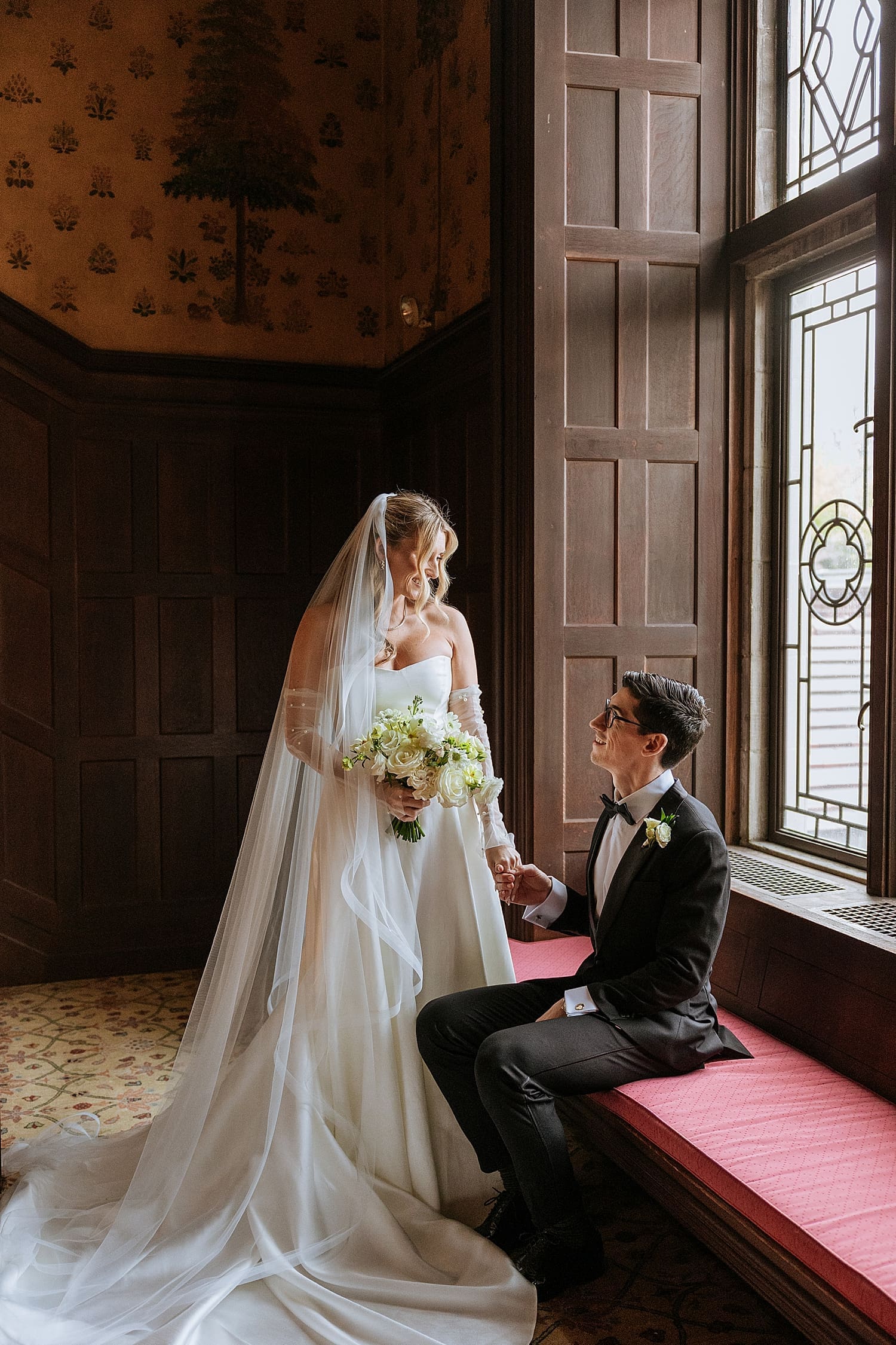 bride looks down at seated groom in window seat at Waveny House